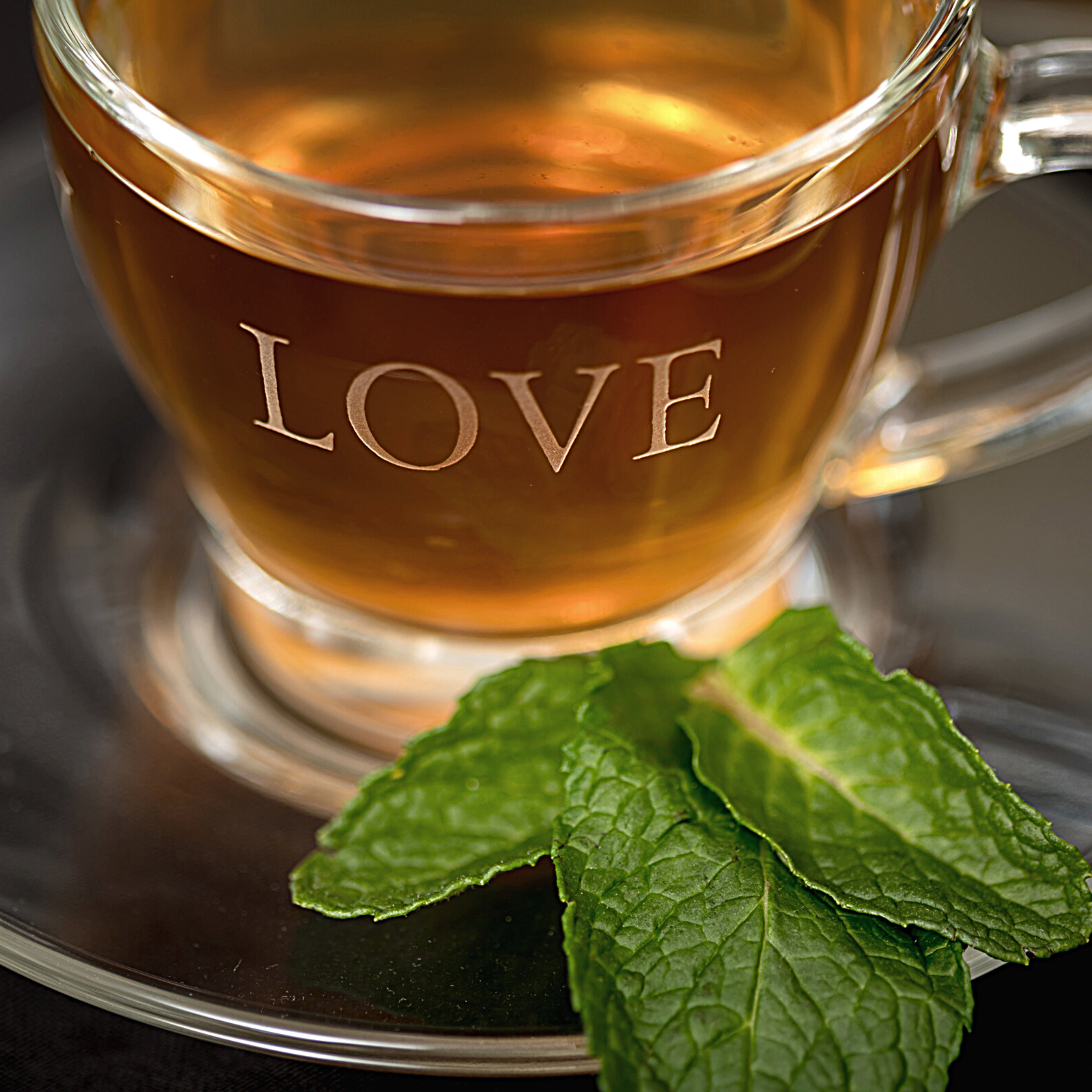 “Glass tea cup etched with the word ‘Love’ filled with herbal tea on a glass saucer”